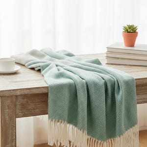 Könnte beinhalten: Hellgrüne und weiße Decke mit Fischgrätmuster und Fransen, drapiert auf einem Holztisch. Eine weiße Tasse mit Untertasse, eine kleine Topfpflanze und Bücher sind ebenfalls zu sehen.