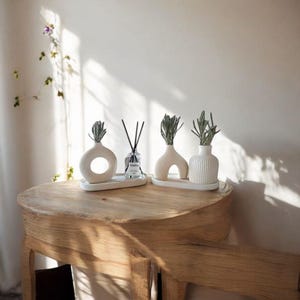 May include: A wooden table displays three white ceramic vases with sprigs of greenery, and a glass diffuser bottle with black reeds. The vases have unique shapes, including a ring and an arch. The arrangement is on a white tray.