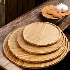 May include: A stack of four round, light brown bamboo serving trays of varying sizes. The trays have a natural wood grain and a raised edge. A smaller tray holds two donuts and a white teapot.