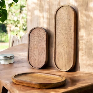 Könnte beinhalten: Drei Holztabletts in verschiedenen Größen. Die Tabletts haben eine ovale Form mit erhöhten Kanten. Das Holz hat eine natürliche Maserung und einen warmen, braunen Farbton. Die Tabletts werden auf einer Holzoberfläche ausgestellt, mit einem Glas im Hintergrund. Die Tabletts eignen sich zum Servieren oder Ausstellen.