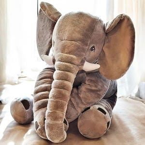 Puede incluir: Un peluche de elefante grande, en tonos grises y marrones. El elefante tiene orejas grandes, una larga trompa y colmillos blancos. Está sentado con las piernas recogidas, y la apariencia general es suave y achuchable.