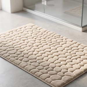 Op de afbeelding: Beige badmat met kiezeltextuur. De rechthoekige mat heeft een patroon van afgeronde, verhoogde vormen, waardoor een 3D-effect ontstaat. De mat ligt op een lichtgrijze vloer, in de buurt van een douche en een kleine houten kruk.