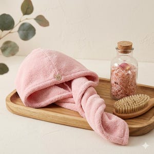 Może przedstawiać: Różowy ręcznik do włosów z mikrofibry z małym guzikiem w kształcie misia. Ręcznik jest złożony i skręcony, spoczywający na drewnianej tacy ze szklanym słoikiem z różową solą do kąpieli i drewnianą szczotką.