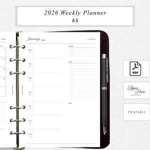 May include: A 2026 weekly planner in A5 size, open to a January page. The planner features a brown leather cover, a black pen, and a white interior with sections for priorities and to-do lists. The image also includes a PDF icon and the text "Paper Dove Designs."