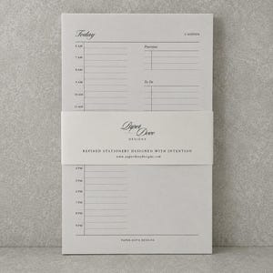 Könnte beinhalten: Ein hellgrauer Tagesplaner-Block mit einem Zeitplan von 6 bis 21 Uhr, Abschnitten für Prioritäten und Aufgaben sowie einem zentralen Papierband mit dem Text Paper Dove Designs, Refined Stationery Designed With Intention.