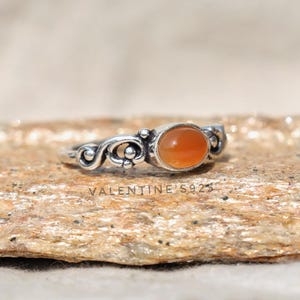 May include: A silver ring with an oval, orange gemstone. The ring features an ornate, swirling design on the band and the text "VALENTINE'S S925". The ring is set on a textured, tan surface.
