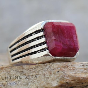 May include: A silver ring with a large, rectangular, deep red gemstone. The ring band features four horizontal black lines. The text "VALENTINE S925" is visible on the ring. The ring is displayed on a piece of wood.