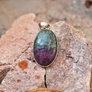 Puede incluir: Un colgante de forma ovalada con una piedra preciosa de rubí zoisita pulida que presenta zonas de color verde y magenta púrpura intenso, engastado en un sencillo bisel de metal plateado con una pequeña anilla integrada. El texto VALENTINES925 es visible.