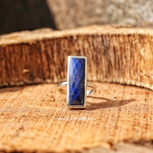 May include: A silver ring featuring a tall, rectangular, faceted lapis lazuli gemstone with a deep blue color and subtle gold flecks. The stone is set in a smooth silver bezel on a simple silver band. The text VALENTINES925 is visible.