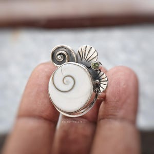 Puede incluir: Un anillo de plata con una concha de ojo de shiva blanca circular con un patrón de espiral natural. El engaste incluye metal plateado decorativo con un motivo de espiral, detalles en forma de abanico y una pequeña piedra verde redonda.
