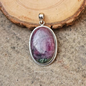 May include: An oval pendant featuring a polished, mottled ruby-zoisite gemstone with deep magenta and dark green inclusions, encased in a simple silver setting with a smooth bail at the top. The text VALENTINES925 is visible at the bottom.