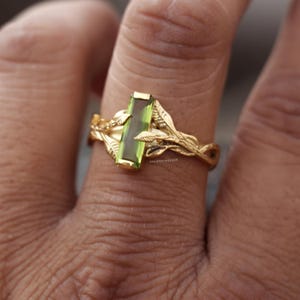 May include: A gold ring featuring a rectangular, vertically oriented green gemstone set within a band designed to resemble intertwined branches and leaves. The small text VALENTINES925 is engraved on the side of the band near the central stone.