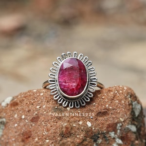 May include: A silver ring featuring a large, oval-shaped, faceted deep pink gemstone set in a decorative scalloped frame. The metal band is thin and polished. The text VALENTINES925 is visible at the base of the ring setting.