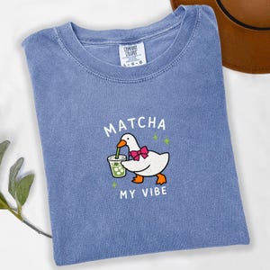 May include: A blue short-sleeve t-shirt featuring a graphic of a white goose wearing a pink bow tie and drinking a green matcha beverage. The text MATCHA MY VIBE is printed in white above and below the goose, accompanied by small green stars.