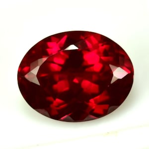 May include: A faceted, oval-shaped, deep red gemstone. The gem has a high degree of clarity, with multiple facets that reflect light, creating a brilliant sparkle. The stone is set against a white background.