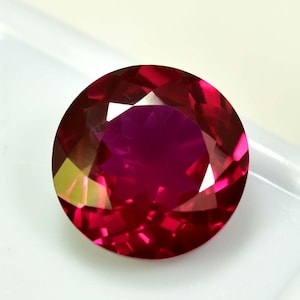 May include: A round-cut ruby gemstone with multiple facets. The gem is a deep red colour with internal reflections, set against a white background. The cut of the stone creates multiple facets that reflect light, giving it a brilliant appearance.