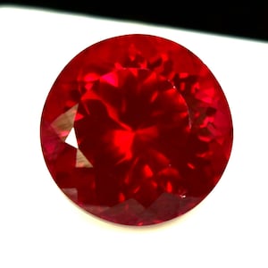 Puede incluir: Una gema redonda, facetada, de color rojo intenso. La gema tiene una superficie brillante y reflectante, con múltiples facetas que capturan y refractan la luz. La piedra está sobre un fondo blanco, resaltando su rico color y claridad. Adecuada para joyería.