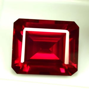 Peut inclure: Une pierre précieuse rouge foncé, taillée en rectangle et bordée de blanc. La gemme a une surface brillante et réfléchissante, mettant en valeur sa structure interne. La pierre est posée sur un fond blanc.