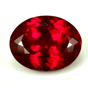 May include: A faceted, oval-cut, deep red gemstone. The gem has a high degree of clarity, with multiple facets that reflect light, creating a brilliant sparkle. The stone is set against a white background.