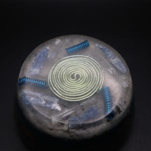 May include: A circular orgonite piece, grey in colour, with blue crystals and three blue springs. A silver spiral sits at the centre. The orgonite is set against a black backdrop.