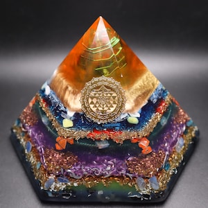 Könnte beinhalten: Eine mehrschichtige Orgonpyramide mit einem goldenen Sri Yantra Symbol. Die Pyramide weist eine Vielzahl von Farben auf, darunter Orange, Gold, Blau, Lila und Grün, mit eingebetteten Kristallen und Kupfer. Ein dekorativer Gegenstand.