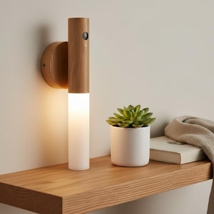 Puede incluir: Lámpara de pared de madera con diseño cilíndrico. La lámpara tiene una sección de vidrio esmerilado que emite una luz cálida. Una pequeña suculenta en una maceta blanca se encuentra en una estantería de madera.