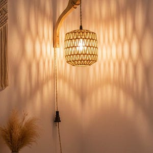 Op de afbeelding: Een geweven rotan hanglamp hangt aan een houten arm die aan de muur is bevestigd. Het licht werpt een patroon van schaduw op de muur. De lamp maakt deel uit van een slaapkamer- of woonkamerdecor.