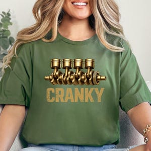May include: An olive green t-shirt with a gold-coloured engine piston design and the word "CRANKY" in a distressed font. The graphic is centered on the shirt, creating a bold statement.