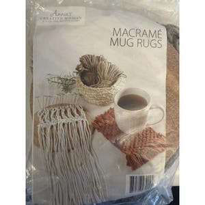 May include: A kit for making macramé mug rugs. The image shows a white mug with a beverage, a finished brown rug, and materials including yarn and a partially completed rug. The text on the package reads "Annie's Creative Woman Kit of the Month Club."