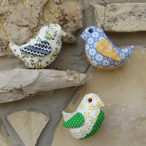 May include: Three fabric bird ornaments with hanging strings. One bird is cream-colored with a navy and white wing pattern. Another is blue with white floral details and a yellow wing. The third is white with green polka dots.