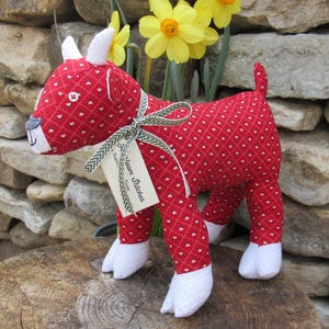 Puede incluir: Un peluche de cabra roja hecho a mano con un patrón de corazones blancos, cuernos blancos y pezuñas blancas. La cabra tiene una cinta verde y blanca atada al cuello y una etiqueta que dice "Naomi Stitches". Los narcisos amarillos están en el fondo.