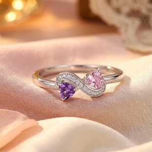 Puede incluir: Un anillo de plata con un diseño de símbolo de infinito. El anillo presenta una piedra preciosa púrpura y rosa en forma de corazón, cada una rodeada de pequeñas piedras transparentes. El anillo está sobre una superficie de satén rosa, con pétalos y flores blancas.