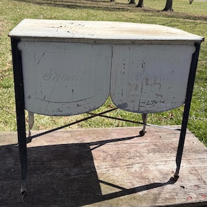May include: An antique, two-basin wash tub with a distressed, off-white finish. The tub has a metal frame with black legs and small wheels. The word "Ideal" is embossed on the front of each basin. The tub sits on a wooden surface.