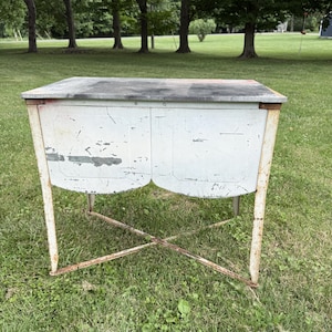 May include: A vintage metal washstand featuring a rectangular gray top and a white cabinet body with two front panels. The structure is supported by four thin legs connected by a cross-brace, showing visible paint wear, rust, and surface distress.