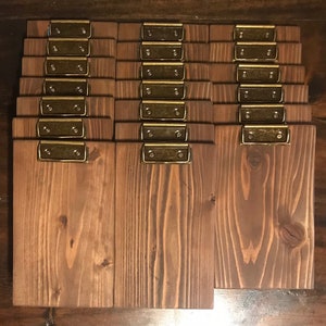 May include: A collection of eighteen rectangular wooden clipboards arranged in three columns. Each clipboard features a medium-brown stained wood grain finish and is equipped with a metallic brass-colored spring-loaded clip at the top edge.