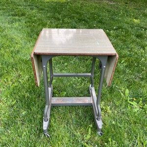 May include: An antique folding table with a light wood grain top and a metal frame. The table has drop leaves and is supported by a gray metal frame with wheels. The table is shown outdoors on a grassy surface.