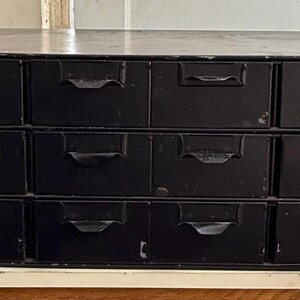May include: A black metal cabinet with fifteen drawers. Each drawer has a curved handle. The cabinet is rectangular and sits on a white surface. The cabinet appears to be vintage or antique.