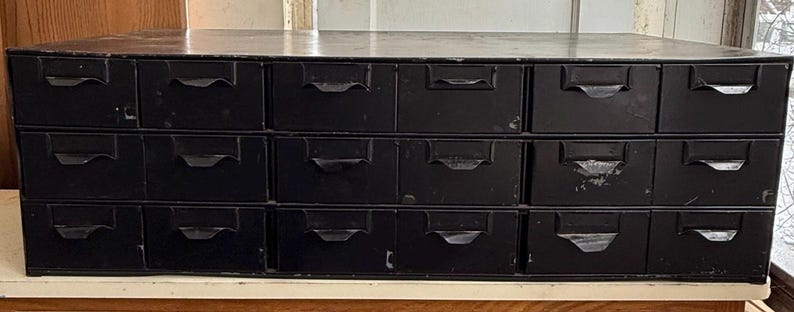 May include: A black metal cabinet with fifteen drawers. Each drawer has a curved handle. The cabinet is rectangular and sits on a white surface. The cabinet appears to be vintage or antique.