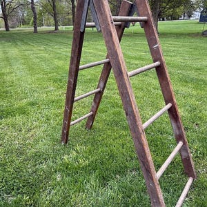 May include: A weathered, wooden A-frame ladder with metal hinges, set on a grassy lawn. The ladder has multiple rungs and a rustic, aged appearance. The background features trees and a glimpse of a building.