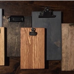 May include: Five wooden clipboards with different colored stains. The clipboards have metal clips in various finishes, including gold, silver, and black.