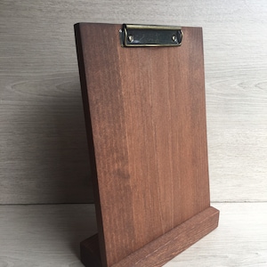 May include: A vertical wooden clipboard stand featuring a dark brown stained finish with a visible wood grain. The board is attached to a rectangular base and includes a metallic, antique-style brass clip mechanism at the top for securing papers.