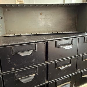 May include: A vintage metal parts cabinet with multiple drawers and a large open top bin. The cabinet is painted black with a distressed finish, and the bin is a light gray color. The drawers have curved metal handles.
