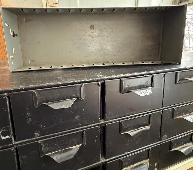 May include: A vintage metal parts cabinet with multiple drawers and a large open top bin. The cabinet is painted black with a distressed finish, and the bin is a light gray color. The drawers have curved metal handles.