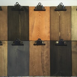 May include: A collection of ten rectangular wooden clipboards arranged in two rows of five. Each board features a distinct wood stain color ranging from light tan to dark charcoal, all equipped with a black metal clip at the top center.