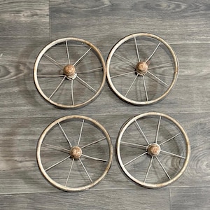 May include: Four antique-style metal wheels with a distressed, off-white finish. Each wheel features a central wooden hub and thin, silver spokes. The wheels are arranged in a square, suggesting a set for a small cart or decorative piece.