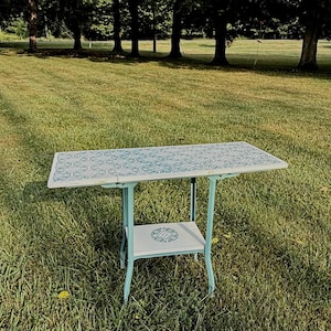 May include: A rectangular folding table with a light blue metal frame and a white tabletop featuring a repeating blue geometric tile pattern. The table includes a lower shelf with a centered circular blue geometric design on a white surface.