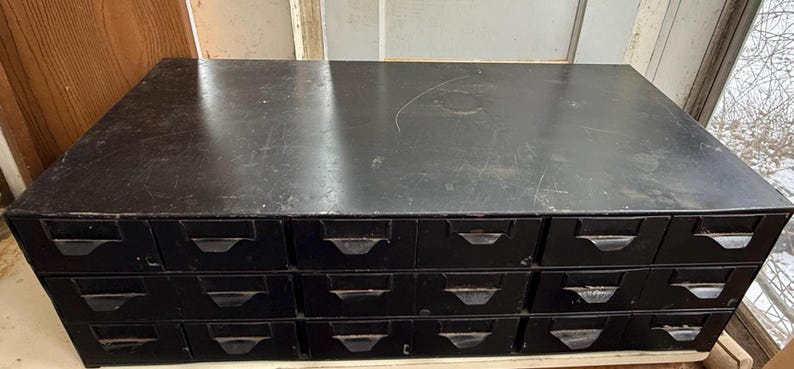 May include: A black, vintage-style cabinet with multiple drawers. The cabinet has a rectangular shape and a flat top. Each drawer has a metal handle. The cabinet appears to be made of wood and has a distressed finish.