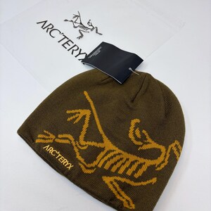 K&ouml;nnte beinhalten: Dunkelolivgr&uuml;ne M&uuml;tze mit goldfarbenem, stilisiertem Dinosaurierskelett-Motiv. Der Markenname "ARC'TERYX" ist goldfarben auf der M&uuml;tze aufgedruckt. Ein schwarzes Etikett ist angebracht.