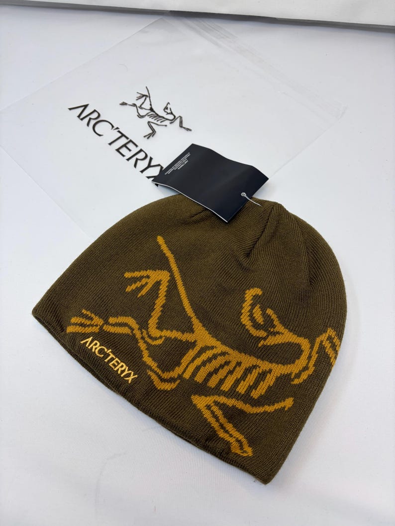K&ouml;nnte beinhalten: Dunkelolivgr&uuml;ne M&uuml;tze mit goldfarbenem, stilisiertem Dinosaurierskelett-Motiv. Der Markenname "ARC'TERYX" ist goldfarben auf der M&uuml;tze aufgedruckt. Ein schwarzes Etikett ist angebracht.