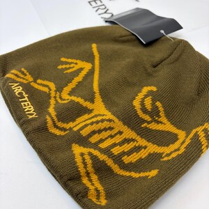 K&ouml;nnte beinhalten: Olivgr&uuml;ne M&uuml;tze mit gelber Grafik eines stilisierten Wesens und dem Wort "ARCTERYX" in Gelb. Ein schwarzes Etikett ist angebracht. Die M&uuml;tze ist aus Strickmaterial.